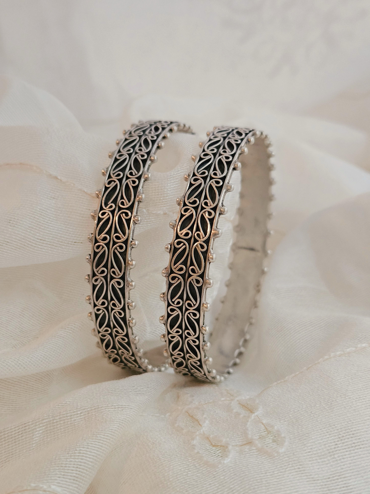 Pair of oxidized silver bangles with paisley filigree scrollwork and beaded edges on ivory fabric