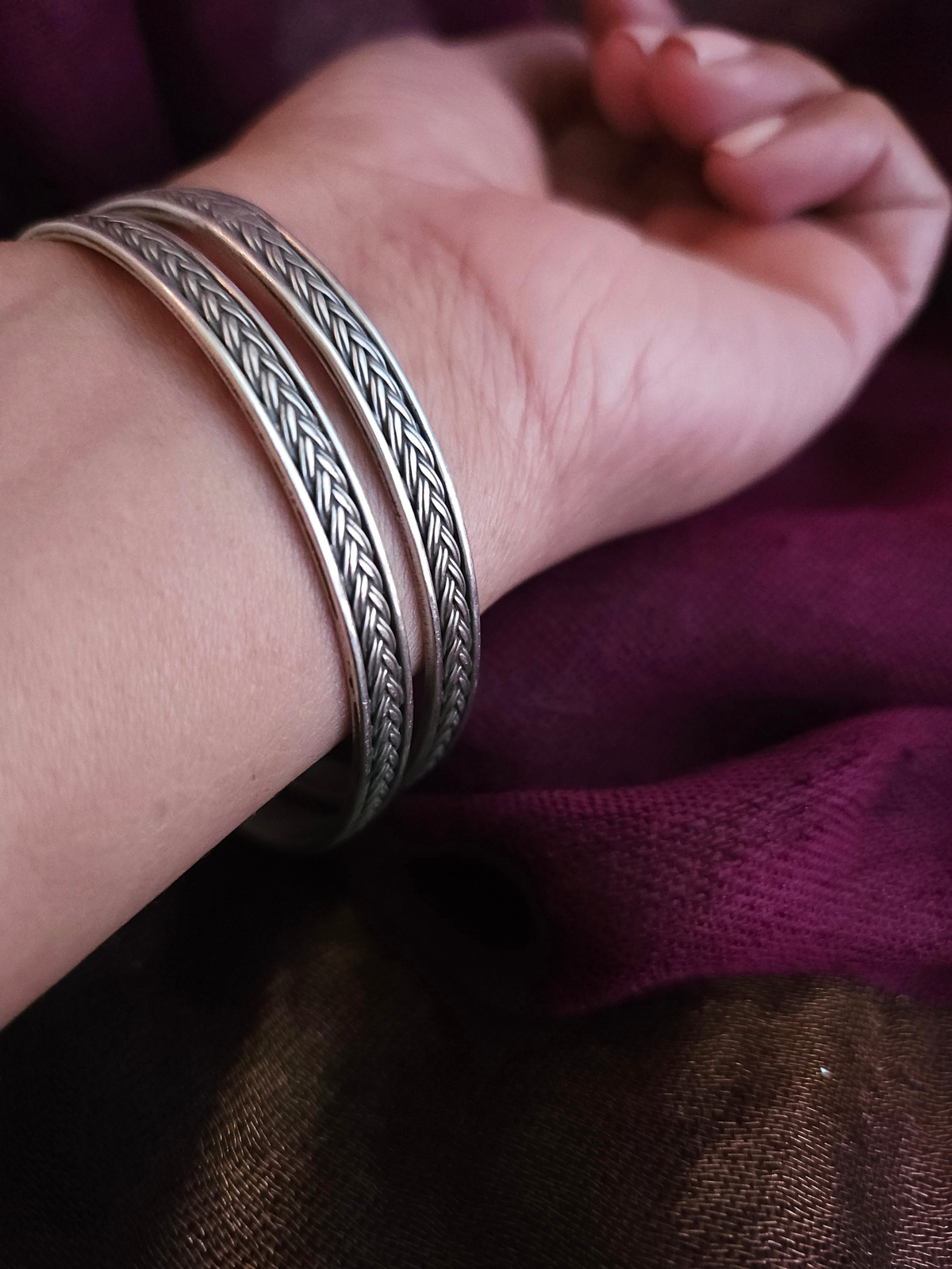 Two oxidized 925 sterling silver braided bangles with polished rims on a wrist against a plum silk saree